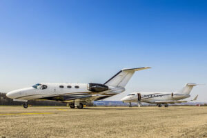 Two private jets on a clear day with a blue sky in the background. The jet in the foreground is white with a blue stripe and has its left wing extended, while the jet in the background is also white and stationary.