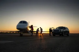 A private jet parked on an airport runway with a car nearby during sunset. Silhouettes of people walking towards the jet are visible against the warm glow of the setting sun in the background. The image has a darkened overlay.